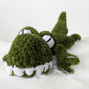 Puede incluir: Un juguete de peluche de cocodrilo verde hecho a mano con dientes blancos y detalles en los ojos. El suave reptil de ganchillo est&aacute; tumbado sobre una superficie blanca y esponjosa, mostrando sus detalles y su dise&ntilde;o tierno.