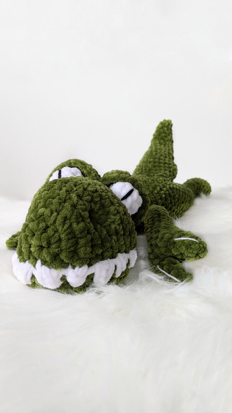 Puede incluir: Un juguete de peluche de cocodrilo verde hecho a mano con dientes blancos y detalles en los ojos. El suave reptil de ganchillo est&aacute; tumbado sobre una superficie blanca y esponjosa, mostrando sus detalles y su dise&ntilde;o tierno.