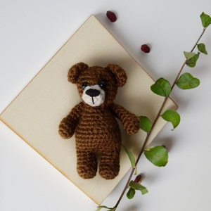 May include: A brown crocheted teddy bear with black eyes and a brown nose. The bear is sitting on a light brown wooden surface.