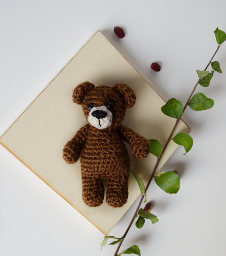May include: A brown crocheted teddy bear with black eyes and a brown nose. The bear is sitting on a light brown wooden surface.