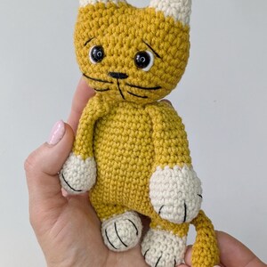 K&ouml;nnte beinhalten: Handgefertigte H&auml;kelkatzenpuppe in Gelb und Creme. Die Katze hat schwarz gestickte Gesichtsz&uuml;ge, einschlie&szlig;lich Augen, Nase und Schnurrhaare. Die Pfoten und Ohren der Puppe sind cremefarben. Dieses weiche Spielzeug ist eine charmante Erg&auml;nzung.