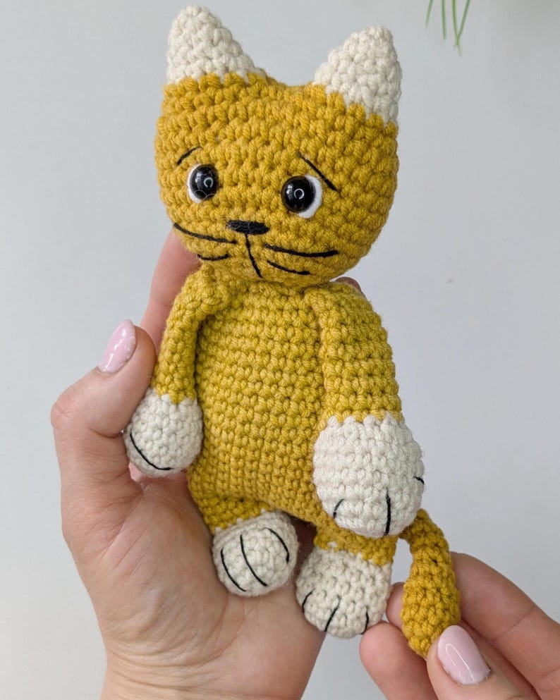 K&ouml;nnte beinhalten: Handgefertigte H&auml;kelkatzenpuppe in Gelb und Creme. Die Katze hat schwarz gestickte Gesichtsz&uuml;ge, einschlie&szlig;lich Augen, Nase und Schnurrhaare. Die Pfoten und Ohren der Puppe sind cremefarben. Dieses weiche Spielzeug ist eine charmante Erg&auml;nzung.