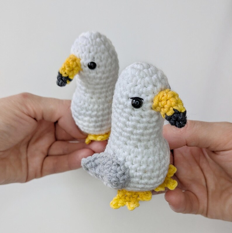 Op de afbeelding: Twee kleine gehaakte meeuwfiguren met witte lichamen, grijze vleugels, gele snavels met zwarte punten en kleine zwarte kraaloogjes. Elke vogel heeft gele zwemvliezen en is gemaakt met een gestructureerd haakpatroon.