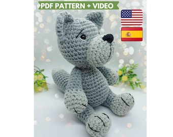 Può includere: Giocattolo di amigurumi a forma di lupo in crochet grigio seduto su una superficie bianca con piante verdi sullo sfondo. Il giocattolo ha occhi neri e un naso nero. Il testo "PDF PATTERN + VIDEO" è nella parte superiore dell'immagine. Le bandiere americana e spagnola sono nell'angolo in alto a destra dell'immagine.