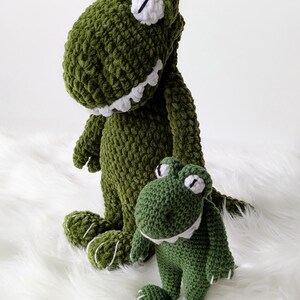 Puede incluir: Dos peluches de dinosaurio verdes tejidos a ganchillo. El dinosaurio m&aacute;s grande est&aacute; de pie y el m&aacute;s peque&ntilde;o est&aacute; sentado. Ambos tienen ojos, dientes y detalles blancos en los pies. Los juguetes est&aacute;n sobre una superficie blanca y esponjosa.