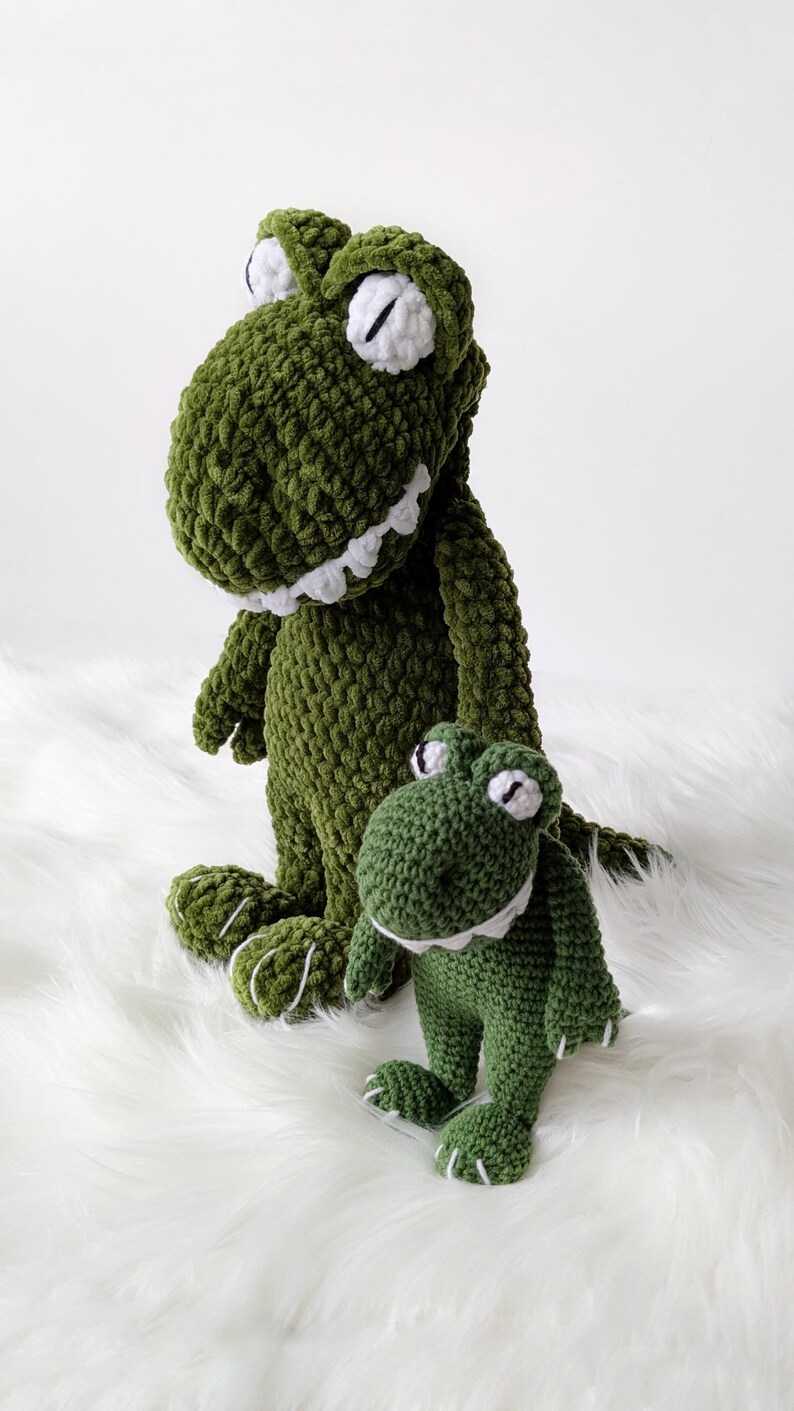 Puede incluir: Dos peluches de dinosaurio verdes tejidos a ganchillo. El dinosaurio m&aacute;s grande est&aacute; de pie y el m&aacute;s peque&ntilde;o est&aacute; sentado. Ambos tienen ojos, dientes y detalles blancos en los pies. Los juguetes est&aacute;n sobre una superficie blanca y esponjosa.