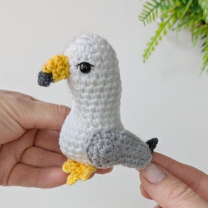 Op de afbeelding: Een kleine, handgemaakte gehaakte meeuwfiguur met een witte kop en lichaam, grijze vleugels, een gele snavel met een donkergrijze punt en gele pootjes. De vogel heeft &eacute;&eacute;n zwart plastic kraaloog aan de zijkant van de kop.