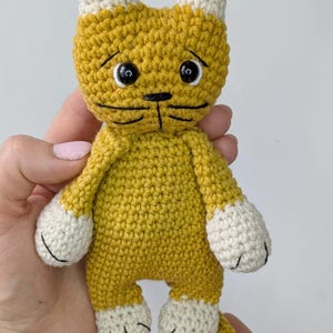K&ouml;nnte beinhalten: Eine handgefertigte H&auml;kelkatzenpuppe in Gelb- und Wei&szlig;t&ouml;nen. Die Katze hat einen traurigen Ausdruck mit schwarzen Stickdetails f&uuml;r Augen, Nase und Mund. Die Ohren, Pfoten und F&uuml;&szlig;e der Katze sind wei&szlig;. Die Puppe ist ein Stofftier.