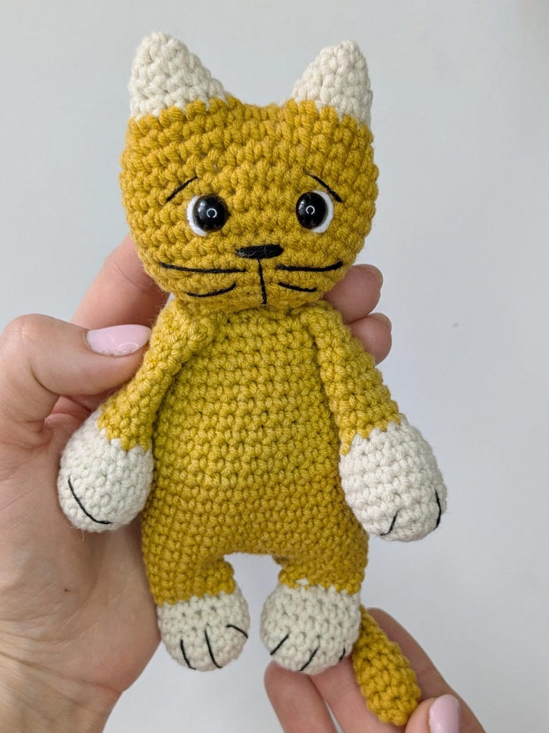 K&ouml;nnte beinhalten: Eine handgefertigte H&auml;kelkatzenpuppe in Gelb- und Wei&szlig;t&ouml;nen. Die Katze hat einen traurigen Ausdruck mit schwarzen Stickdetails f&uuml;r Augen, Nase und Mund. Die Ohren, Pfoten und F&uuml;&szlig;e der Katze sind wei&szlig;. Die Puppe ist ein Stofftier.