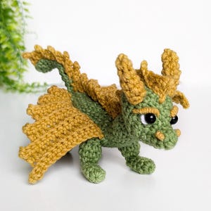 May include: A green and gold crocheted dragon toy with black eyes. The dragon has a long tail and wings.