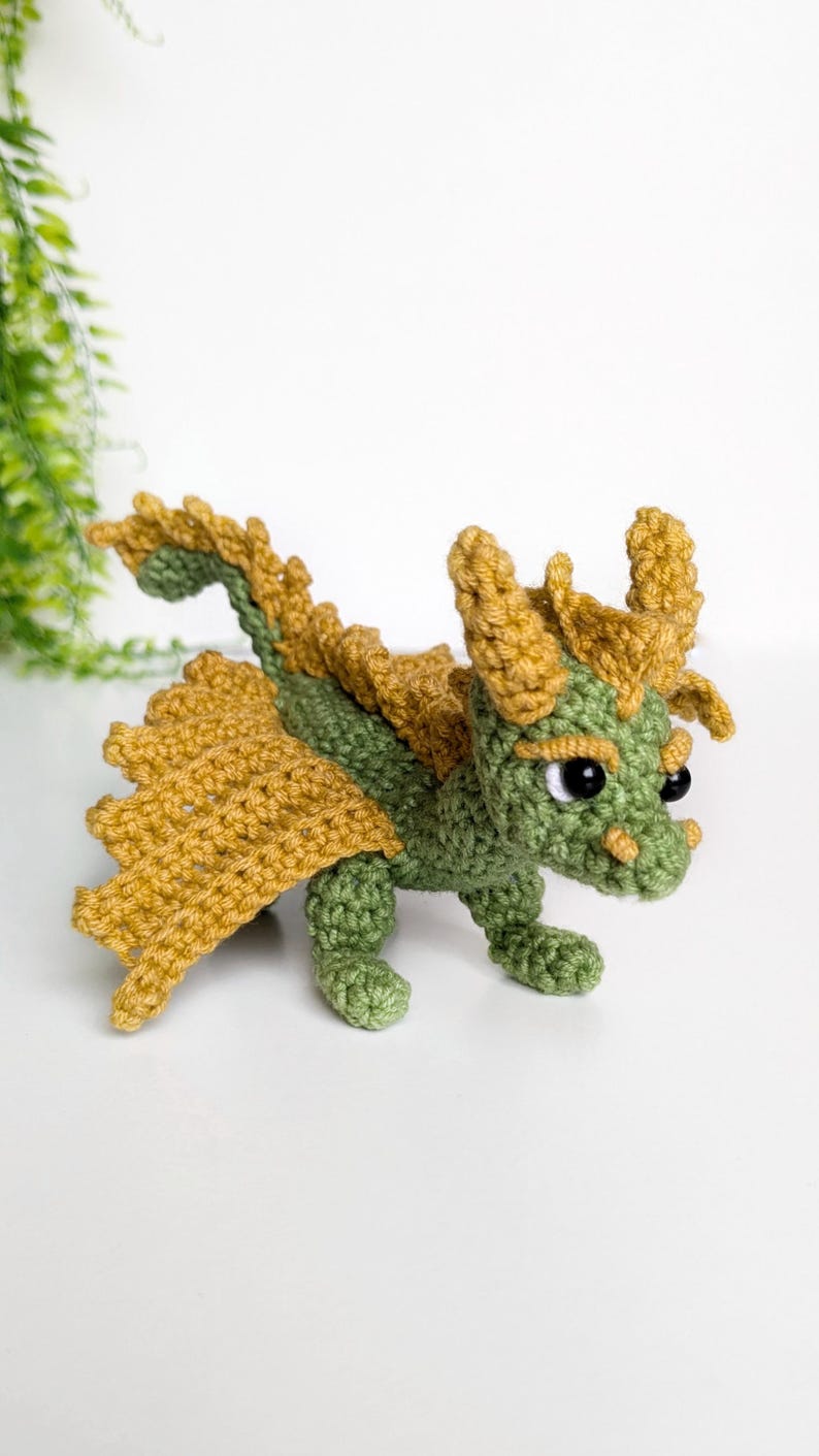 May include: A green and gold crocheted dragon toy with black eyes. The dragon has a long tail and wings.
