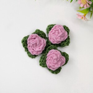 May include: Three handmade crochet flowers. Each flower features a pink rose atop a green leaf base. The flowers are arranged on a white surface, with green foliage and pink flowers in the background.