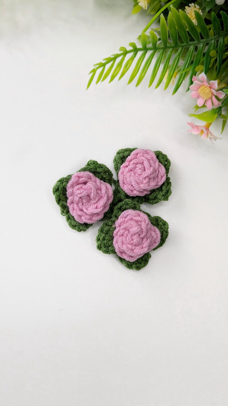 May include: Three handmade crochet flowers. Each flower features a pink rose atop a green leaf base. The flowers are arranged on a white surface, with green foliage and pink flowers in the background.