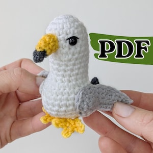 Op de afbeelding: Een klein, handgemaakt gehaakt meeuwspeeltje met een witte kop en lichaam, grijze vleugels, een gele snavel met een donkergrijze punt en gele pootjes. Het speeltje heeft een zwart kraaloog en een groene grafische overlay met de tekst PDF.