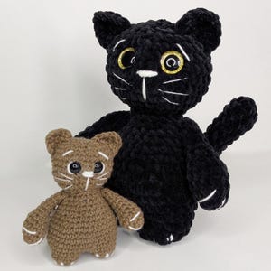 May include: Two handmade crochet cat plushies, one larger black cat with glittery gold eyes and one smaller brown cat. Both feature white embroidered whiskers, a white nose detail, and white stitched accents on their paws against a plain background.