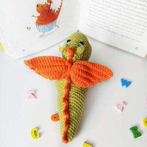 May include: A green crocheted dragon with orange wings and a long tail. The dragon is lying on a white surface with colorful wooden letters scattered around it.