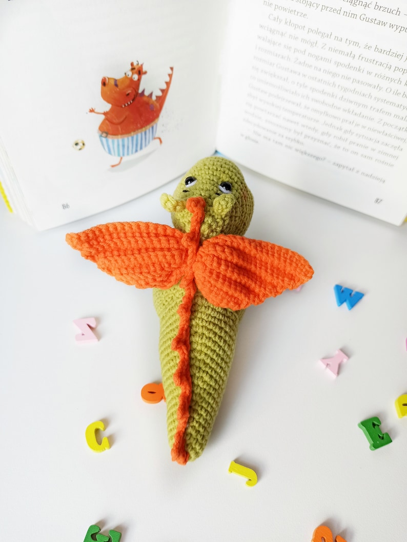 May include: A green crocheted dragon with orange wings and a long tail. The dragon is lying on a white surface with colorful wooden letters scattered around it.