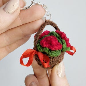 May include: A miniature crochet basket keychain filled with red roses and green leaves. The brown basket has a handle and is tied with a red ribbon. The keychain is attached to a silver ring.