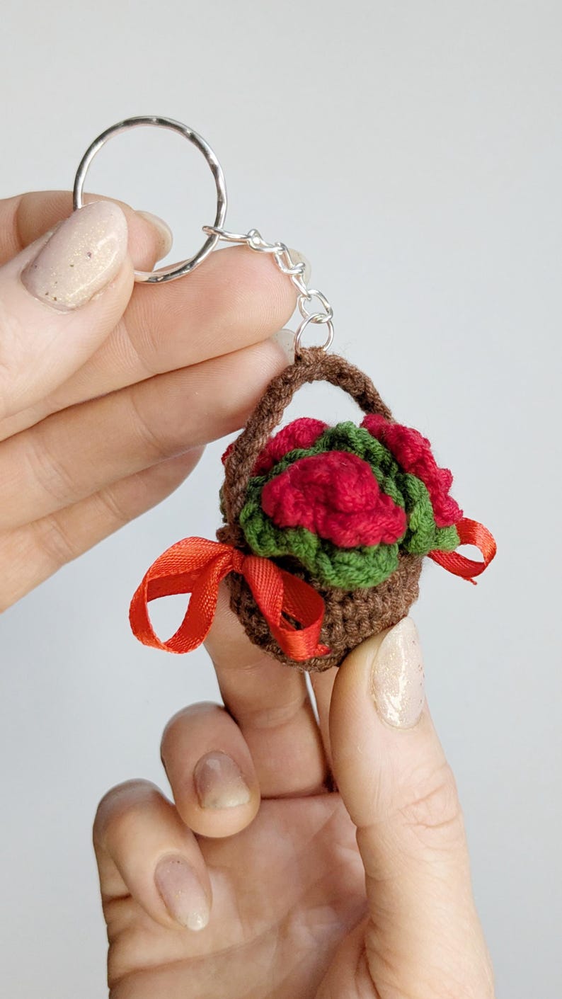 May include: A miniature crochet basket keychain filled with red roses and green leaves. The brown basket has a handle and is tied with a red ribbon. The keychain is attached to a silver ring.
