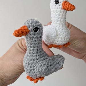 May include: Two small crocheted duck figures, one crafted from light gray yarn and the other from white yarn. Both ducks feature bright orange crocheted beaks and feet, along with small, round, black plastic safety eyes for a detailed appearance.