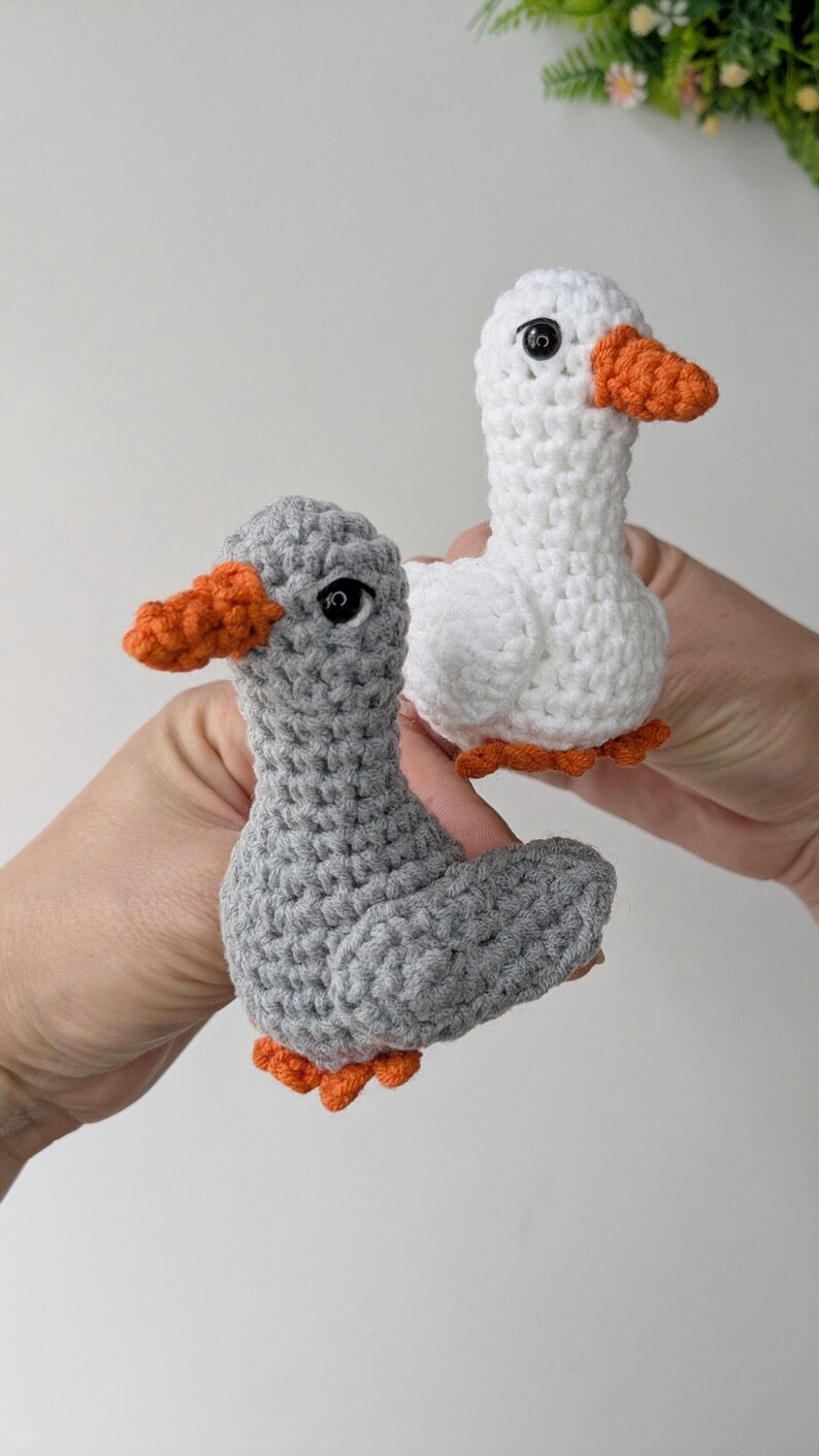 May include: Two small crocheted duck figures, one crafted from light gray yarn and the other from white yarn. Both ducks feature bright orange crocheted beaks and feet, along with small, round, black plastic safety eyes for a detailed appearance.