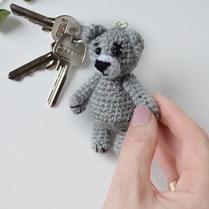 May include: A gray crocheted teddy bear keychain with black eyes and a white nose. The bear is being held by a hand with a gold ring.