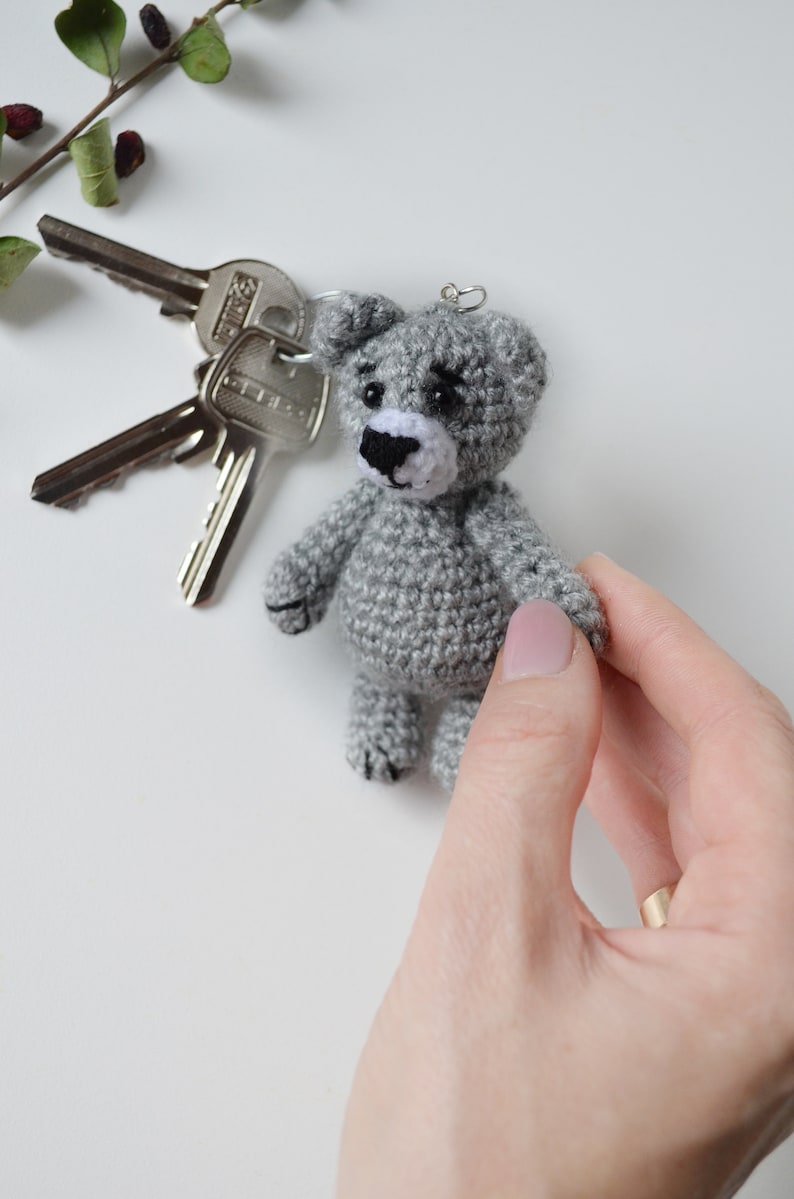 May include: A gray crocheted teddy bear keychain with black eyes and a white nose. The bear is being held by a hand with a gold ring.