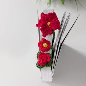 Puede incluir: Un marcap&aacute;ginas de ganchillo con tres flores rojas de centro amarillo y hojas verdes unidas a un tallo verde. El marcap&aacute;ginas est&aacute; colocado verticalmente entre las p&aacute;ginas de un libro blanco, con el dise&ntilde;o floral a lo largo del lomo.