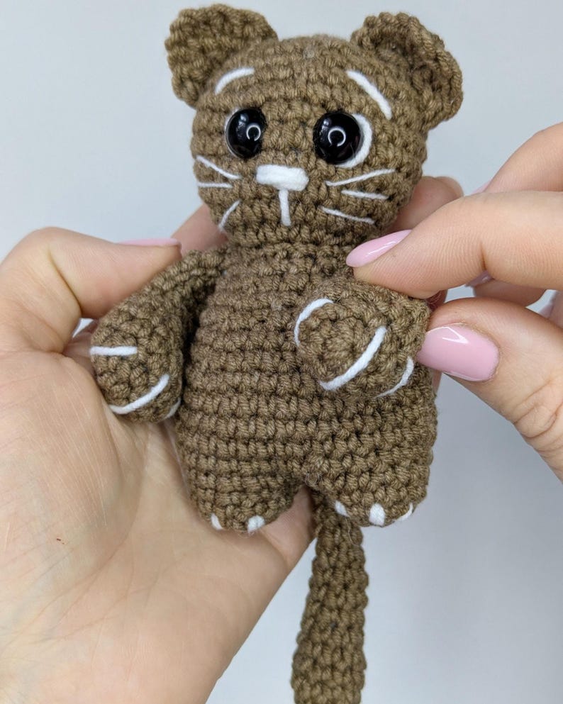 May include: A small brown crocheted cat toy featuring large black plastic safety eyes, white embroidered whiskers, white eyebrow accents, and white stitched details on the paws and tail. The yarn is a medium brown shade with a textured finish.