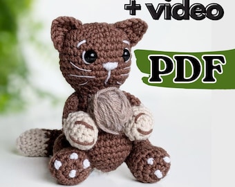 May include: Brown crocheted cat amigurumi holding a ball of yarn. The cat has white accents on its paws, nose, and eyes. The image includes the text " + video PDF".