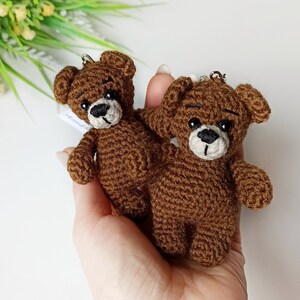 May include: Two brown crocheted teddy bear keychains with black eyes and white noses. The bears are held in a hand.
