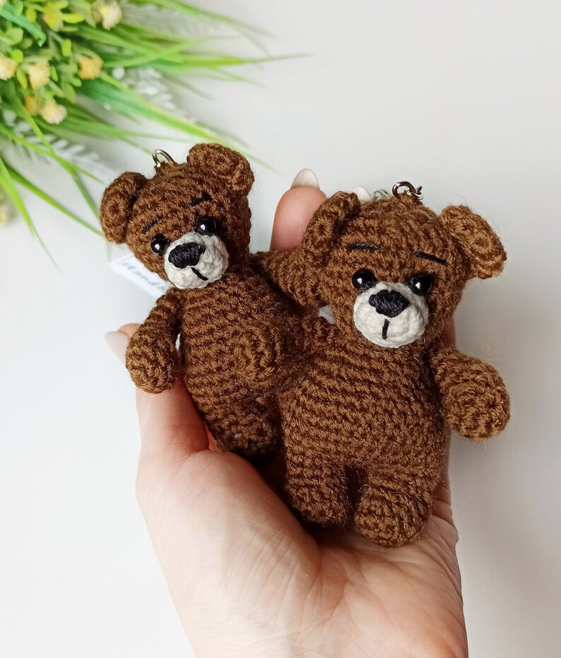 May include: Two brown crocheted teddy bear keychains with black eyes and white noses. The bears are held in a hand.