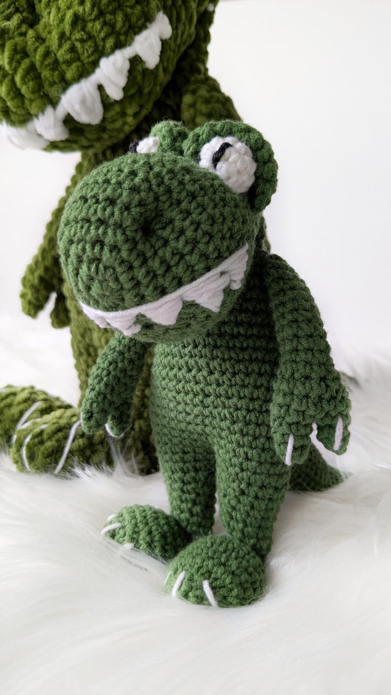Puede incluir: Dos juguetes de dinosaurio de ganchillo verde hechos a mano. Los dinosaurios tienen dientes, ojos y garras blancos. Un dinosaurio est&aacute; en primer plano y otro en segundo plano. Los juguetes est&aacute;n sobre una superficie blanca y esponjosa.