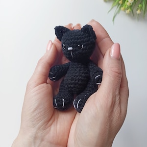 May include: A small black crocheted cat toy with white accents on the paws and face. The cat is being held in the palms of two hands.