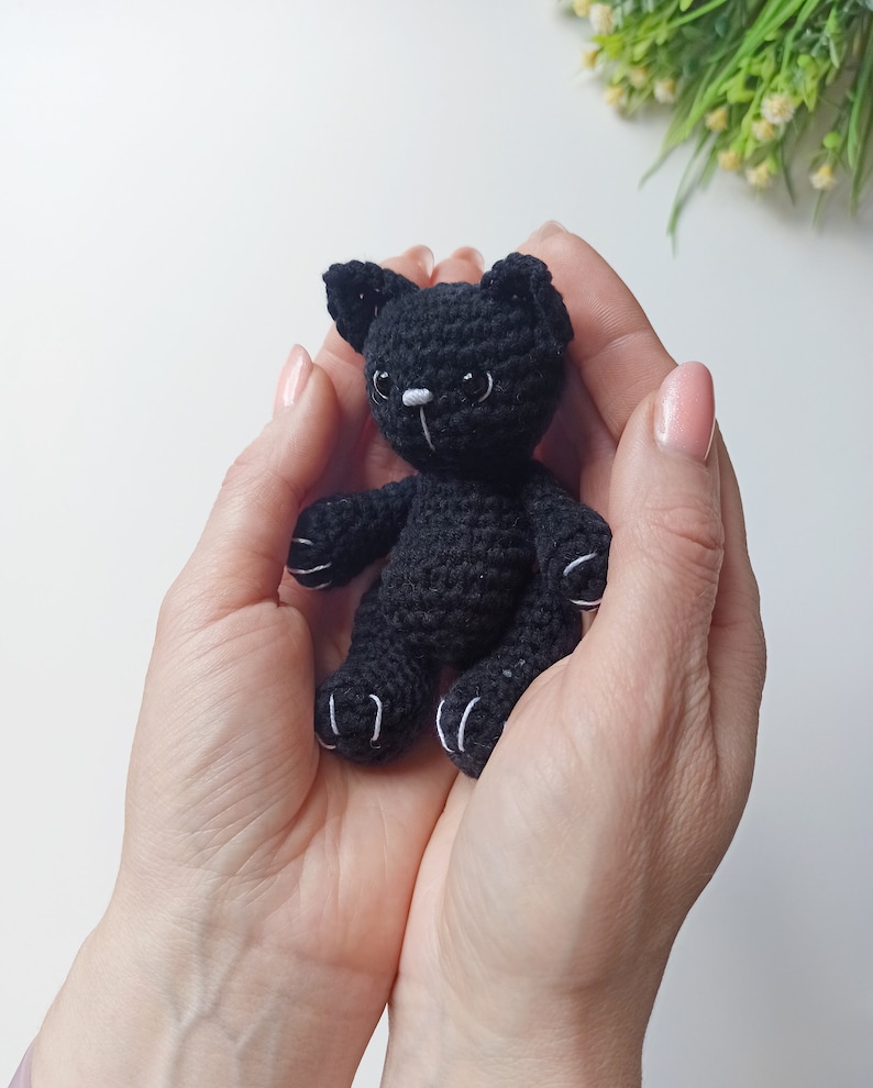 May include: A small black crocheted cat toy with white accents on the paws and face. The cat is being held in the palms of two hands.