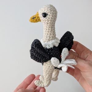 May include: A crocheted vulture toy featuring a cream-colored head and legs, a yellow beak, a black body with dark wings, and a white ruff around the neck. The small stuffed animal has black safety eyes and a white tail detail on the back.