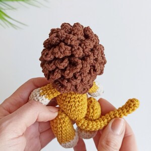 May include: A crocheted lion toy with a brown mane, yellow body, and white feet. The lion is held in two hands.