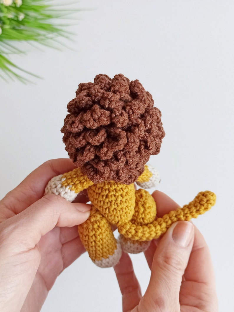 May include: A crocheted lion toy with a brown mane, yellow body, and white feet. The lion is held in two hands.