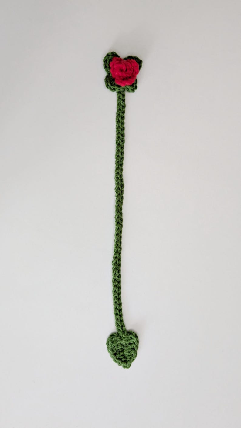 Puede incluir: Un marcap&aacute;ginas de ganchillo largo y fino en hilo de color verde oscuro. La parte superior presenta una peque&ntilde;a rosa roja vibrante tejida a ganchillo rodeada de hojas verdes. El tallo largo y trenzado termina en una hoja verde en forma de coraz&oacute;n.