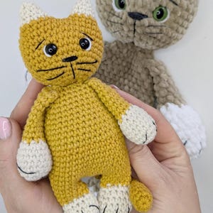 K&ouml;nnte beinhalten: Handgeh&auml;kelte Katzenpuppe in Senfgelb mit wei&szlig;en Akzenten an Pfoten und Ohren. Die Katze hat schwarze Schnurrhaare und einen traurigen Gesichtsausdruck. Eine weitere &auml;hnliche Katze in Beige im Hintergrund.