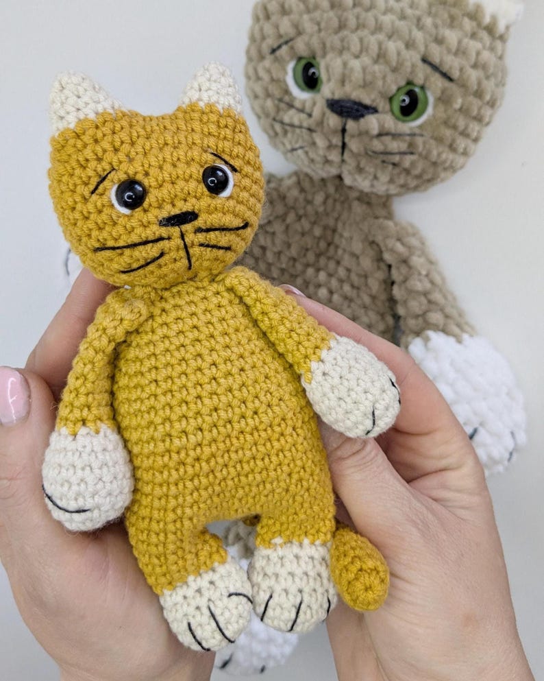 K&ouml;nnte beinhalten: Handgeh&auml;kelte Katzenpuppe in Senfgelb mit wei&szlig;en Akzenten an Pfoten und Ohren. Die Katze hat schwarze Schnurrhaare und einen traurigen Gesichtsausdruck. Eine weitere &auml;hnliche Katze in Beige im Hintergrund.