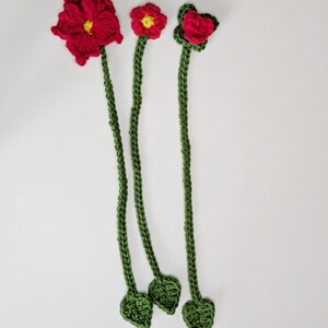 Puede incluir: Tres marcap&aacute;ginas de flores tejidos a mano en ganchillo, con tallos largos de color verde y peque&ntilde;as hojas en la base. Cada marcap&aacute;ginas tiene una flor roja en la parte superior, dos con centros amarillos y una con p&eacute;talos superpuestos.