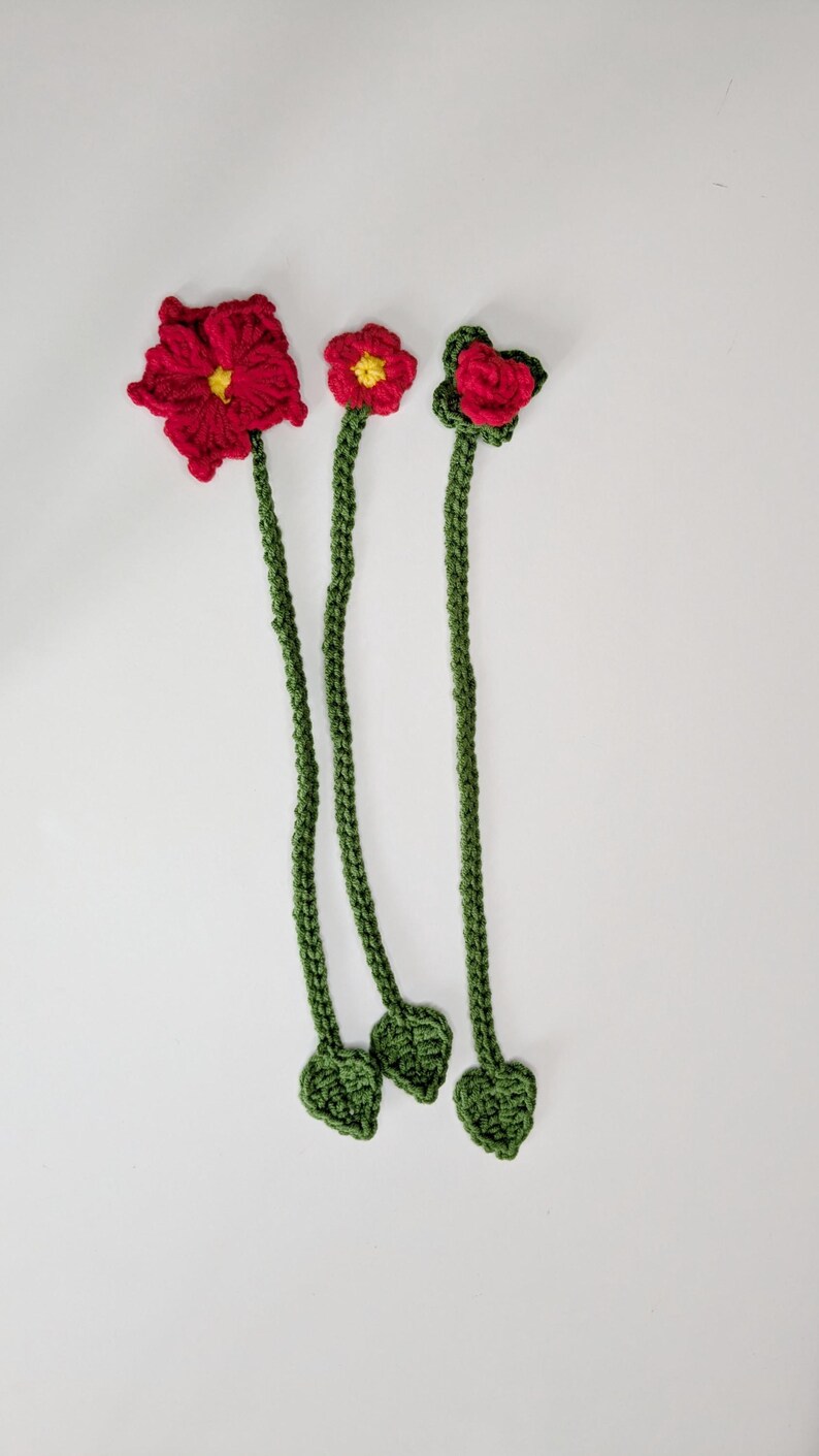 Puede incluir: Tres marcap&aacute;ginas de flores tejidos a mano en ganchillo, con tallos largos de color verde y peque&ntilde;as hojas en la base. Cada marcap&aacute;ginas tiene una flor roja en la parte superior, dos con centros amarillos y una con p&eacute;talos superpuestos.