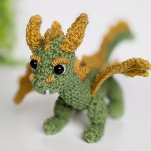 May include: A small green crocheted dragon with yellow horns and wings. The dragon has black eyes and a toothy grin.