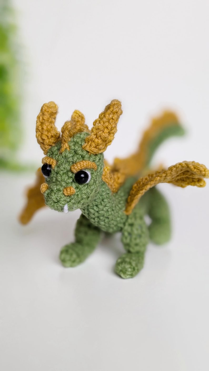 May include: A small green crocheted dragon with yellow horns and wings. The dragon has black eyes and a toothy grin.