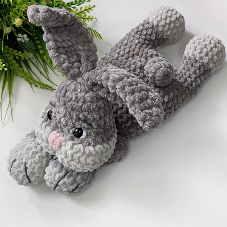 Op de afbeelding: Een handgemaakt grijs gehaakt konijn knuffel met een roze neus en zwarte ogen. Het konijn ligt op zijn rug met zijn armen en benen uitgestrekt. De knuffel is gemaakt van zacht, getextureerd garen.