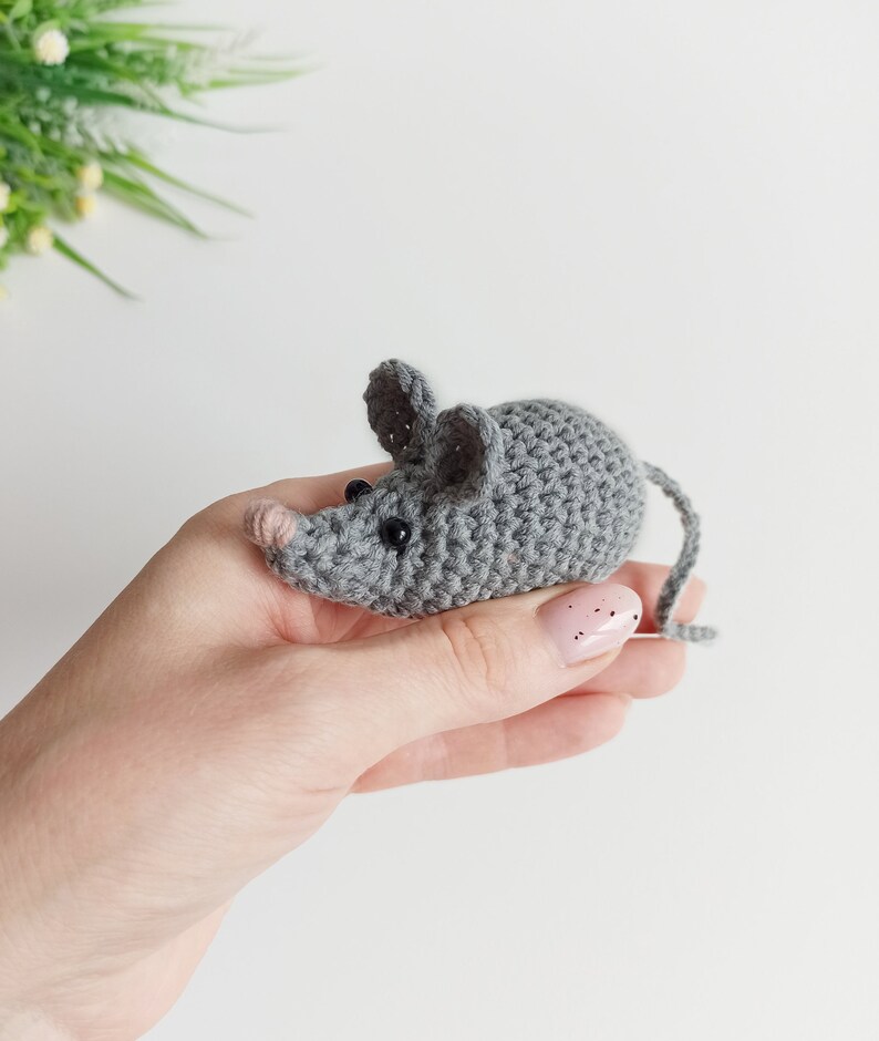 May include: A gray crocheted mouse toy with black eyes. The mouse is being held in a hand with pink nail polish.