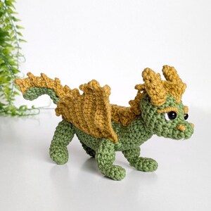 May include: A green and gold crocheted dragon toy with a spiky tail and wings. The dragon has black eyes and a small, toothy mouth.
