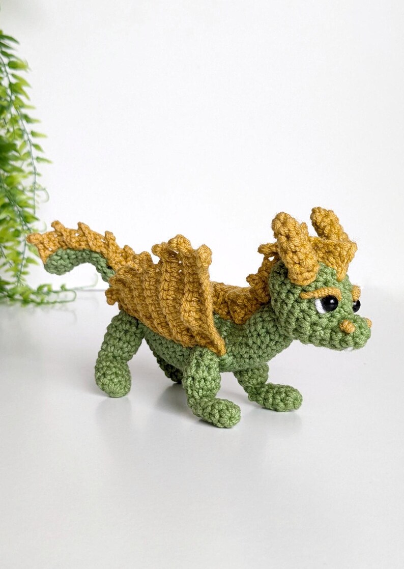 May include: A green and gold crocheted dragon toy with a spiky tail and wings. The dragon has black eyes and a small, toothy mouth.