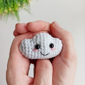 May include: A white crocheted cloud with a smiling face, black eyes, and pink cheeks. The cloud is being held in a person's hands.